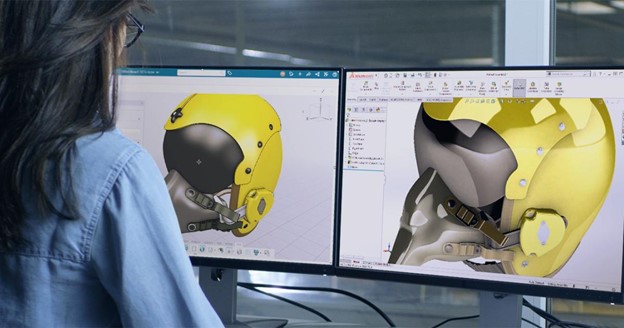 A designer working on a dual-monitor setup, modeling a detailed yellow helmet in SOLIDWORKS and the 3DEXPERIENCE platform.